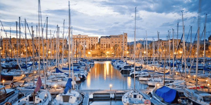 Luxury yachts and sailboats moored in Barcelona harbor at twilight with city lights