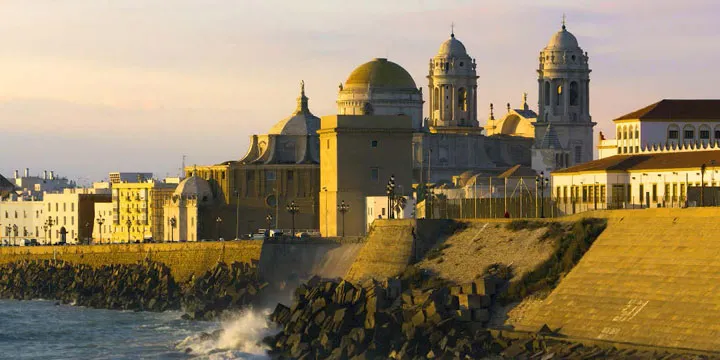 A wide scenic view of the Cádiz coastline in Spain at sunset, featuring the historic Cathedral with its golden dome and white stone towers against a soft evening sky.
