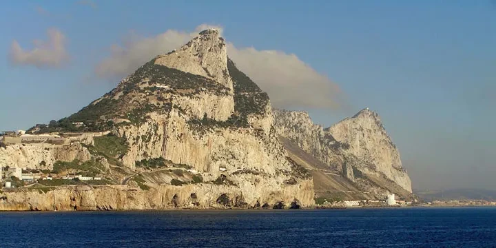 A scenic view of the massive limestone Rock of Gibraltar rising from the blue waters of the Mediterranean under a clear sky.