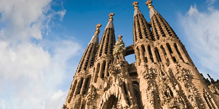 A low-angle view of the intricate stone spires and sculptural details of the Sagrada Família basilica in Barcelona against a blue sky with soft clouds.
